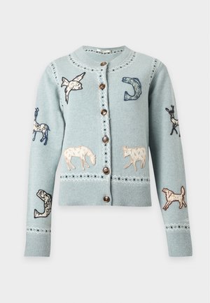 Vrouw die een lichtblauw vest draagt met dierenapplicaties, een blauwe denimrok en zwarte veter-sandalen staand tegen een effen witte achtergrond.