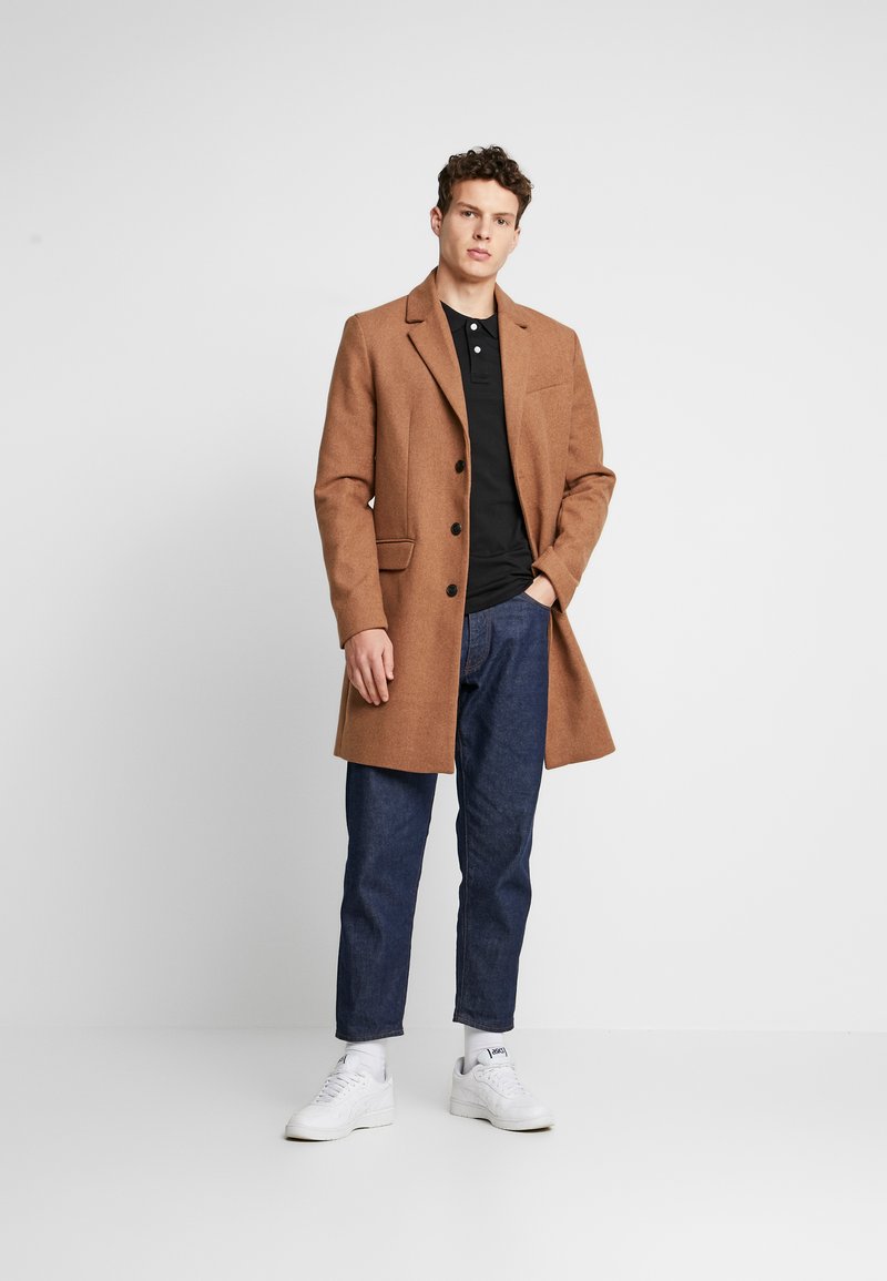 Manteau en mélange de laine marron avec une coupe ajustée, revers crantés, deux poches avant et boutons noirs, associé à un polo noir et un jean bleu.