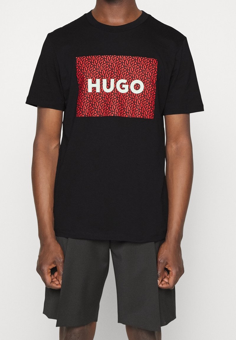 T-shirt en coton noir avec un graphique rectangulaire rouge portant le texte "HUGO" en blanc et un motif répétitif. Design à manches courtes et col rond.