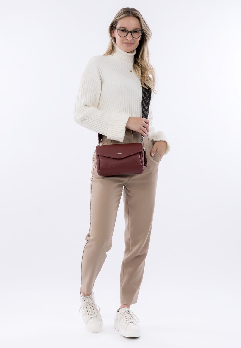 Rote texturierte Leder-Crossbody-Tasche mit einem Magnetverschluss, kombiniert mit einem weißen gerippten Rollkragenpullover und beigen eng anliegenden Hosen.