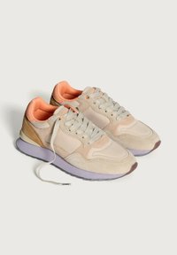 Un paio di sneaker beige e pesca con lacci grigi chiari e suole intermedie viola su uno sfondo bianco.