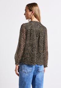 Blusa trasparente marrone con stampa leopardata, caratterizzata da maniche lunghe e dettagli arricciati sul retro, abbinata a jeans azzurri.