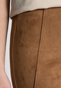 Jupe en daim marron avec une texture douce, présentant des lignes épurées et une finition lisse, s'étendant jusqu'à la taille avec un subtil détail de couture.