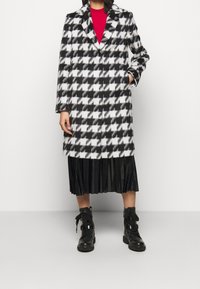 Sort-hvid houndstooth-frakke med hakket krave, enkel knaplukning og lange ærmer, båret over en plisseret sort nederdel og støvler.