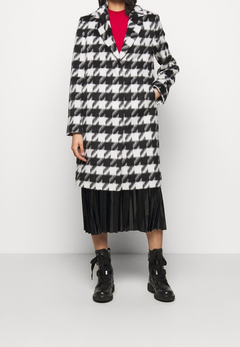 Sort-hvid houndstooth-frakke med hakket krave, enkel knaplukning og lange ærmer, båret over en plisseret sort nederdel og støvler.