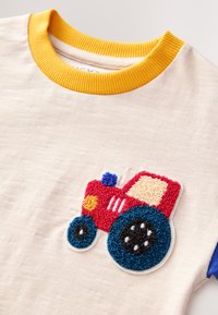 Maglietta beige chiaro con un design colorato di un trattore ricamato in rilievo. Colletto a costine giallo e maniche blu per un contrasto accattivante.