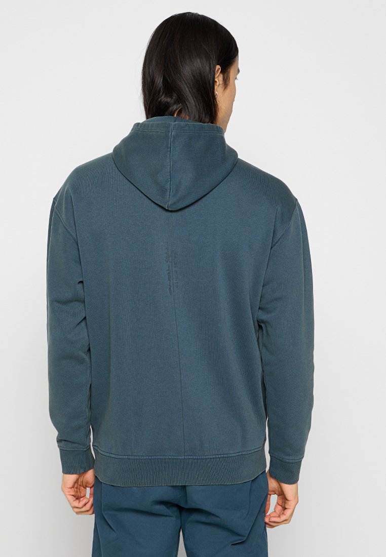 Kaptrowa bluza w ciemnym odcieniu zieleni, o prążkowanej fakturze, z kieszenią typu kangaroo i elastycznymi mankietami, z widocznymi detalami szwów z tyłu.