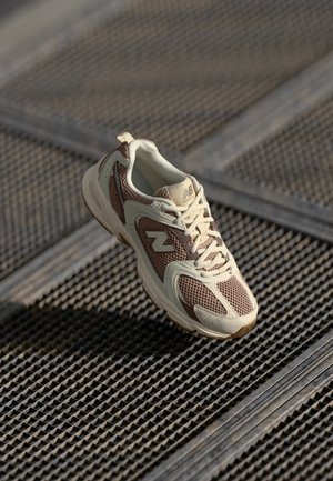 Beige Laufschuh mit Mesh- und synthetischen Einsätzen, ausgestattet mit einem dunkelbraunen Akzent, strukturiertem Sohlenprofil und einem dezenten Logo an der Seite.