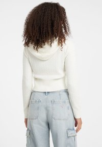 Femeie cu păr creț, îmbrăcată cu un pulover alb cu glugă din tricot cu model și pantaloni cargo din denim ușor bleu, cu talie înaltă, văzută din spate.