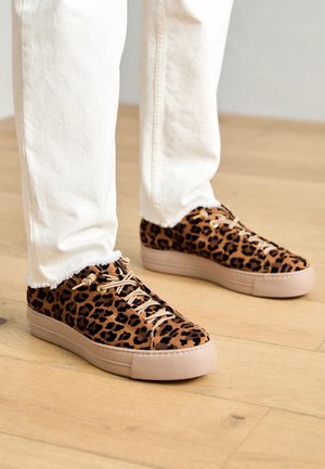 Sneakers med leopardprint, beige såler og snørebånd, båret med off-white bukser med frynset kant på et trægulv.