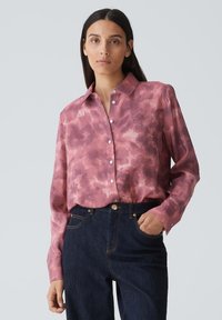 Chemise button-up rose clair avec un motif tie-dye, manches longues et col classique, associée à un jean en denim bleu foncé.
