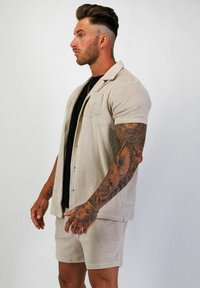 Beige skjorta med kort ärm, krage och framficka, i kombination med matchande shorts. Tyget verkar ha en texturerad yta med en mjuk, avslappnad design.