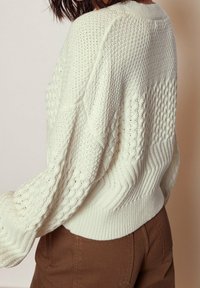 Chunkig, krämfärgad stickad tröja med strukturerade mönster, ribbade ärmslut och en avslappnad passform. Observera den detaljerade kabelstickningsdesignen.