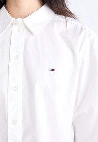 Camisa de algodón blanca con un cuello clásico, botones frontales y un pequeño logo bordado en rojo, blanco y azul en el lado izquierdo del pecho.