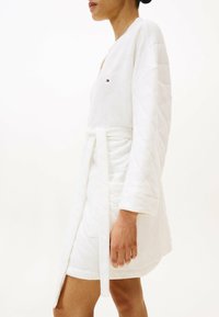 Gewatteerde witte badjas met lange mouwen, ceintuur in de taille en een klein logo op de borst. Glad textuur met een subtiel patroon.