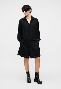 Muž v přehnaně velké černé košili, širokých černých šortkách, černých kotníkových botách z lakované kůže a velkých černých slunečních brýlích, stojící s rukama v kapsách.