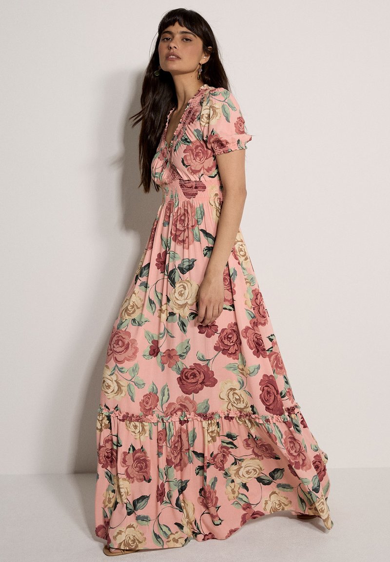 Femme en robe maxi rose à motif floral avec manches courtes bouffantes, encolure à volants et taille froncée, debout devant un fond neutre.