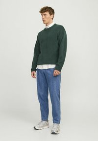 Suéter verde texturizado con cuello redondo, superpuesto a una camisa blanca con cuello, combinado con jeans azules y zapatillas ligeras.