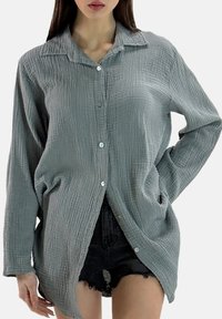 Lichtgrijs, gestructureerd overhemd met knopen, een kraag en lange mouwen. Heeft een losse pasvorm en zijsplitten voor meer bewegingsvrijheid.