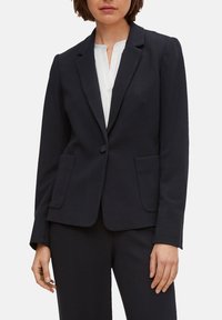 Blazer bleu marine su misura in tessuto texturizzato con chiusura a un bottone, rever a punta e due tasche frontali. Abbinato a una camicia bianca.