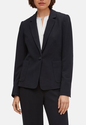 Blazer azul marino ajustado en tejido texturizado con un cierre de un solo botón, solapas con muesca y dos bolsillos frontales. Combinado con una blusa blanca.