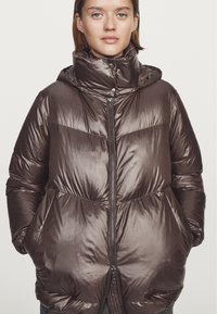 Brązowa puchowa kurtka z wysokim kołnierzem, zamek błyskawiczny i dużymi kieszeniami. Satynowe wykończenie, lekko błyszcząca tekstura oraz poziome przeszycia.