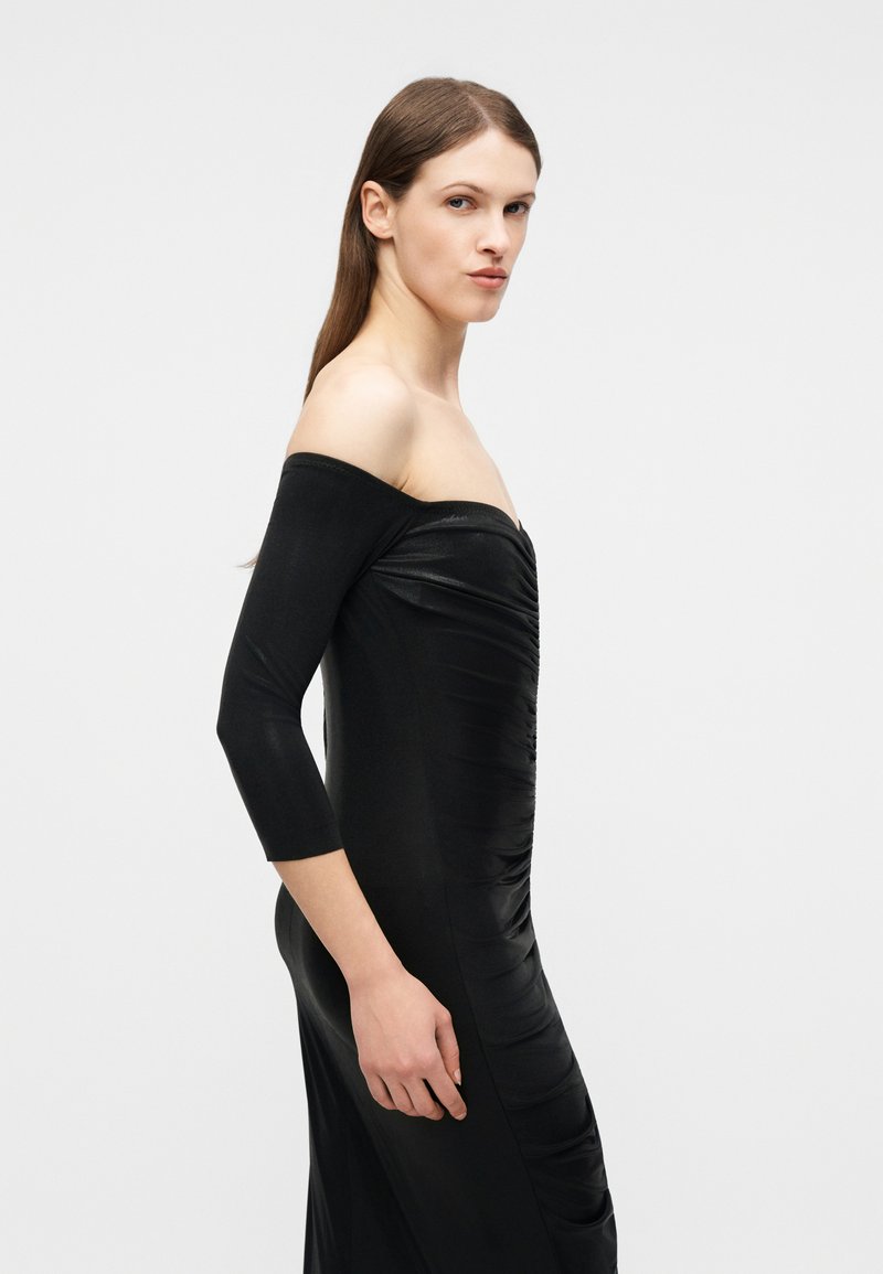 Woman with straight brown hair wearing a fitted, off-the-shoulder black dress with three-quarter sleeves, standing against a plain background.