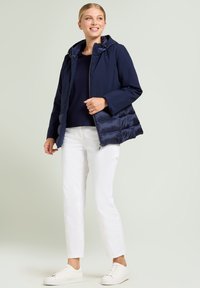 Marineblauw jack met opgeblazen zijpanelen en een capuchon, gecombineerd met een donker shirt en witte broek. Model draagt witte sneakers.