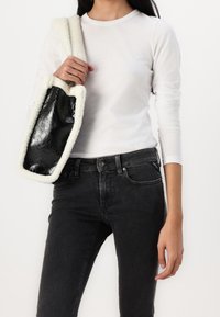 Bolso de imitación de cuero negro con un borde de color crema esponjoso. El modelo lleva una camisa blanca de manga larga y jeans oscuros, mostrando los detalles del bolso.