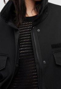 Zwarte jas met een hoge kraag, kliksluitingen en zijzakken, gedragen over een gebreide zwarte top met een opengewerkt patroon. Textuur van de stofdetails zichtbaar.