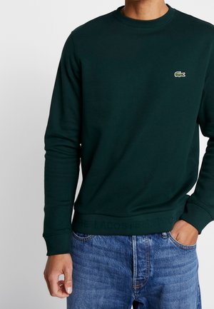 Mężczyzna w ciemnozielonej bluzie Lacoste z logo na piersi i niebieskich dżinsach, z jedną ręką w kieszeni, na jednolitym jasnym tle.