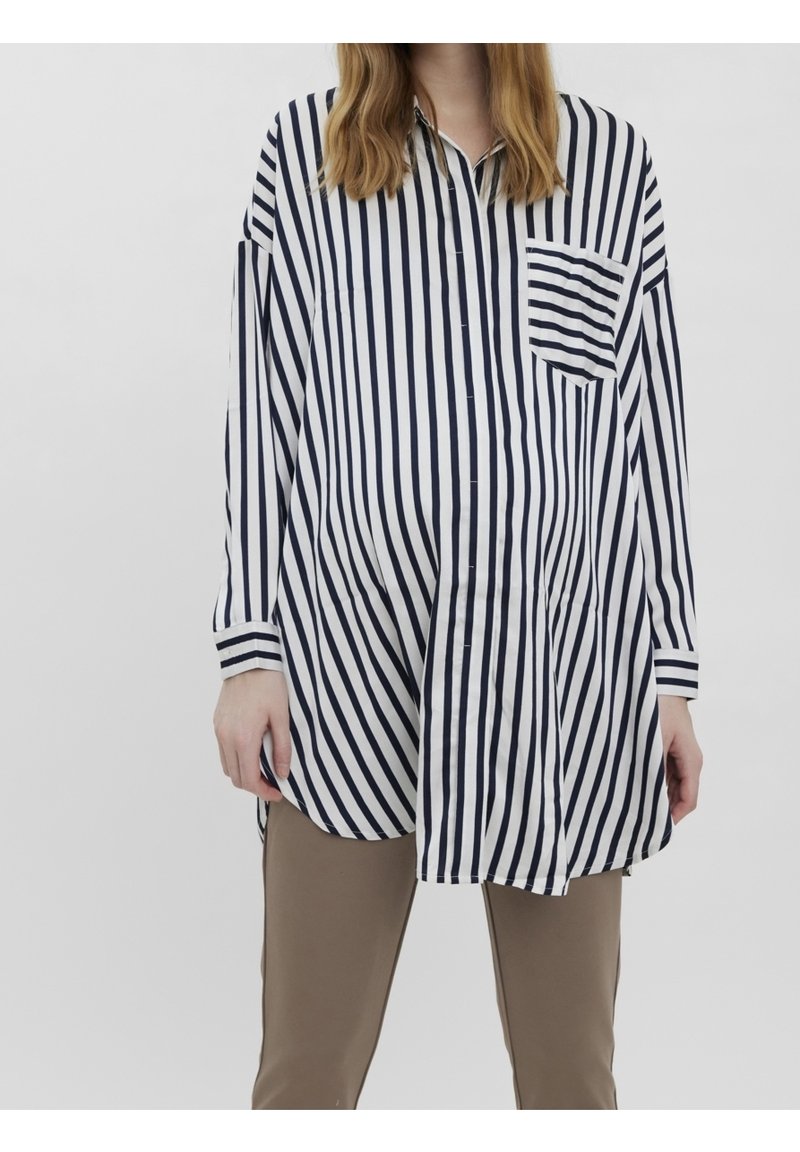Femme portant une chemise blanche à manches longues avec des rayures verticales noires et une poche poitrine rayée, associée à un pantalon beige.