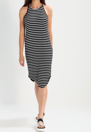 Femme marchant, portant une robe midi sans manches rayée horizontalement noir et blanc et des sandales noires avec des semelles blanches.