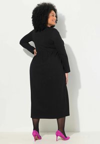 Robe noire à manches longues avec une taille froncée, associée à des talons hauts roses vif. Tissu lisse et silhouette élégante.
