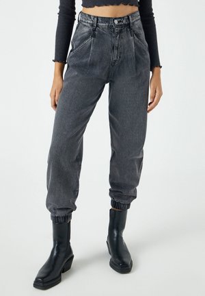 Vrouw draagt high-waisted, zwart gewassen denim joggerbroek met elastische boorden en zwarte enkellaarzen met een vierkante neus.