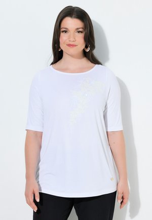 Kobieta o długich ciemnych włosach, ubrana w białą koszulę z krótkim rękawem z delikatnym kwiatowym haftem i czarne spodnie, stojąca na jasnym tle.