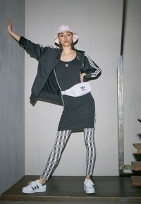 Černé sportovní šaty vrstvené s černou sportovní bundou, bílým opaskem s logem, vzorovanými legínami a bílými teniskami s černými akcenty.