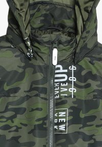 Kamuflage pufferjakke i nuancer af grøn, med en frontlynlås, hætte og trykte hvide tekstaccenter. Glat tekstur, blank overflade.