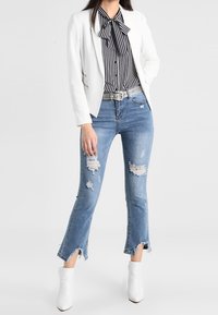 Blazer blanc porté sur un chemisier à col noeud rayé noir et blanc, associé à un jean bleu clair déchiré et à des bottines blanches.