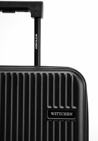 Valigia rigida nera con texture a coste verticali, dotata di manico estraibile e una placca con il logo "WITTCHEN."