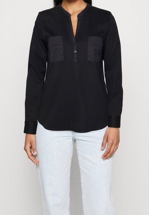 Woman wearing a black long-sleeve blouse with two front chest pockets and light blue jeans, standing against a plain background.