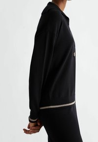 Czarny sweter z kołnierzykiem, wyposażony w zamek błyskawiczny z złotymi akcentami oraz teksturowany dół w złote paski. Miękka tkanina, luźny krój.