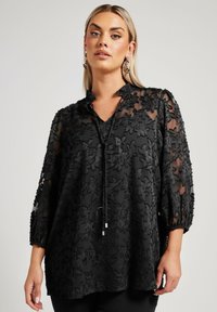 Tunique noire en tissu transparent, ornée d'une broderie florale, avec un col en V et des manches bouffantes trois-quarts. Accentuée par un détail de cordon.