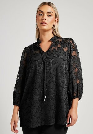 Black tunic top made of sheer fabric, featuring floral embroidery, a V-neck, and three-quarter puff sleeves. Accented with a drawstring detail.