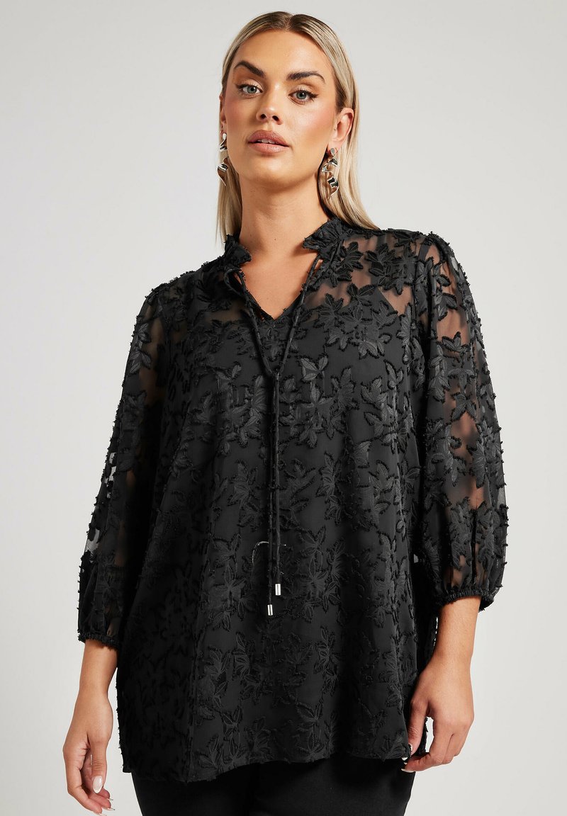 Tunique noire en tissu transparent, ornée d'une broderie florale, avec un col en V et des manches bouffantes trois-quarts. Accentuée par un détail de cordon.