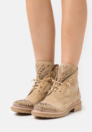 Beige ruskind ankelstøvler med guld- og sølvnitter, snørelukning foran og dekorative guldøjeletter, båret på bare ben.
