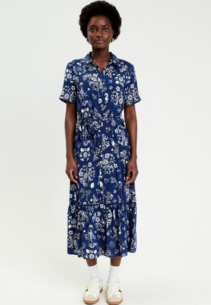 Frau steht und trägt ein blaues, geblümtes Midikleid mit kurzen Ärmeln und weißen Sneakers vor einem einfarbigen Hintergrund.