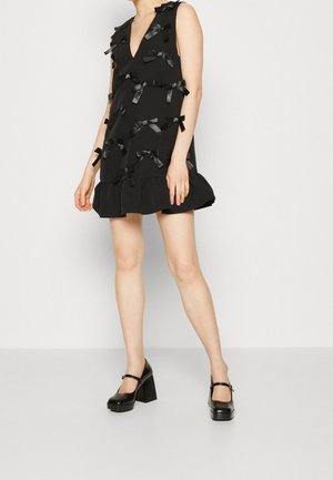 Czarna sukienka bez rękawów z dekoltem w kształcie litery V, ozdobiona wieloma czarnymi wstążkami i falbaną na dole. Modelka nosi czarne buty na słupku.