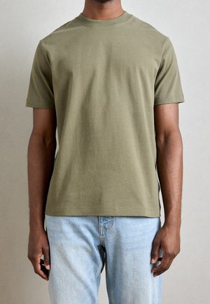 Mężczyzna ubrany w oliwkowo zielony t-shirt z krótkim rękawem i okrągłym dekoltem oraz jasnoniebieskie dżinsy na jednolitym tle.