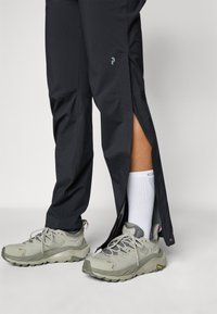 Black lightweight pants with side zippers paired with grey sneakers. Pants feature a sleek design and a subtle logo near the knee.